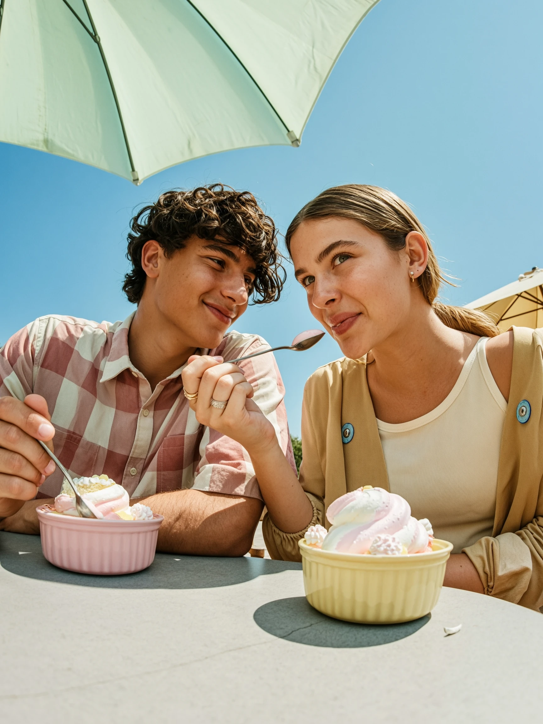 two young people eating dessert together, close-up shot, wide angle lens, exaggerated perspective, sitting at an outdoor table, feeding each other with spoons, playful expressions, summer vibe, bright sunlight, pastel umbrellas above, blue sky, casual candid moment, lifestyle photography, vibrant colors, high contrast, natural skin texture, modern editorial style, high detail