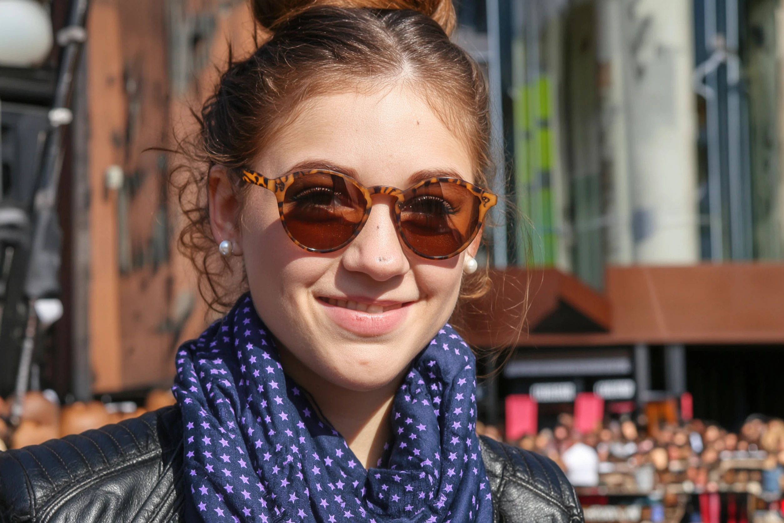 Generate an image that matches the exquisite silhouette outlined in Figure 1, and follows the following description: A young woman smiling on a sunny day, wearing a pair of brown round sunglasses with leopard print patterns on the frames. Her hair is neatly tied up, she wears pearl earrings, a dark blue scarf with purple star patterns around her neck, and a black leather jacket.